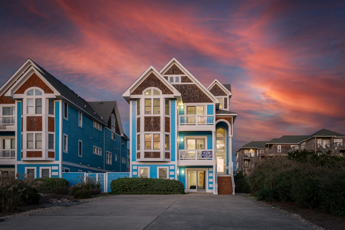The vibrant exterior of the oceanfront home is illuminated by a colorful sunset sky. The structure features multiple levels and large windows, with a welcoming entrance flanked by decorative gables. Lush landscaping surrounds the driveway, adding to the inviting atmosphere.