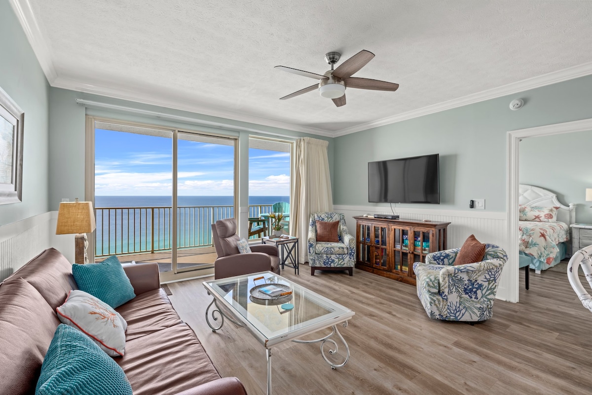 A spacious living area features a brown sofa and two patterned chairs, complemented by a glass coffee table. Large sliding doors offer a view of the ocean and access to a balcony, while a flat-screen television is mounted on the wall.