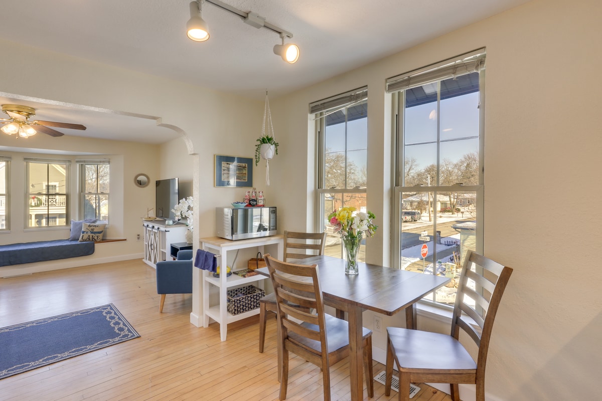 Natural light fills the open living space, showcasing a dining area with a wooden table and chairs. Large windows provide views of the surroundings. A seating area features a daybed, while a TV is mounted on the wall, adding to the welcoming atmosphere.