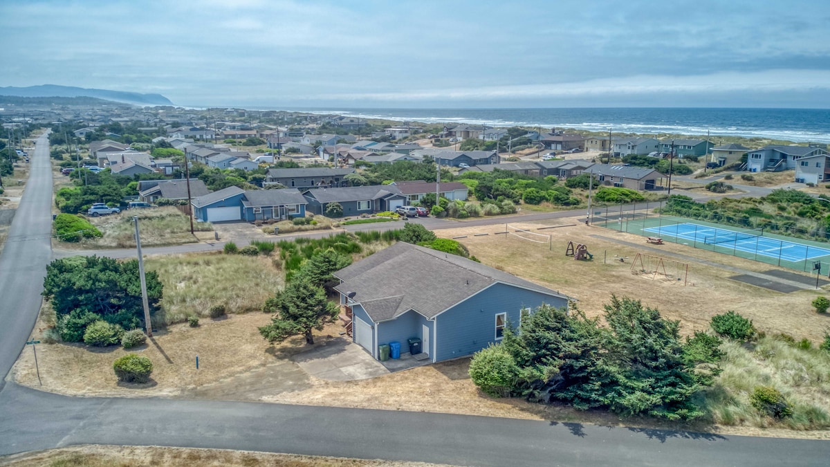 An aerial view of a coastal neighborhood is presented, showcasing the ocean in the distance. A single-level home is visible, with the neighborhood playground and tennis courts nearby. The landscape features a mix of grass and shrubs, with paved roads weaving through the area.