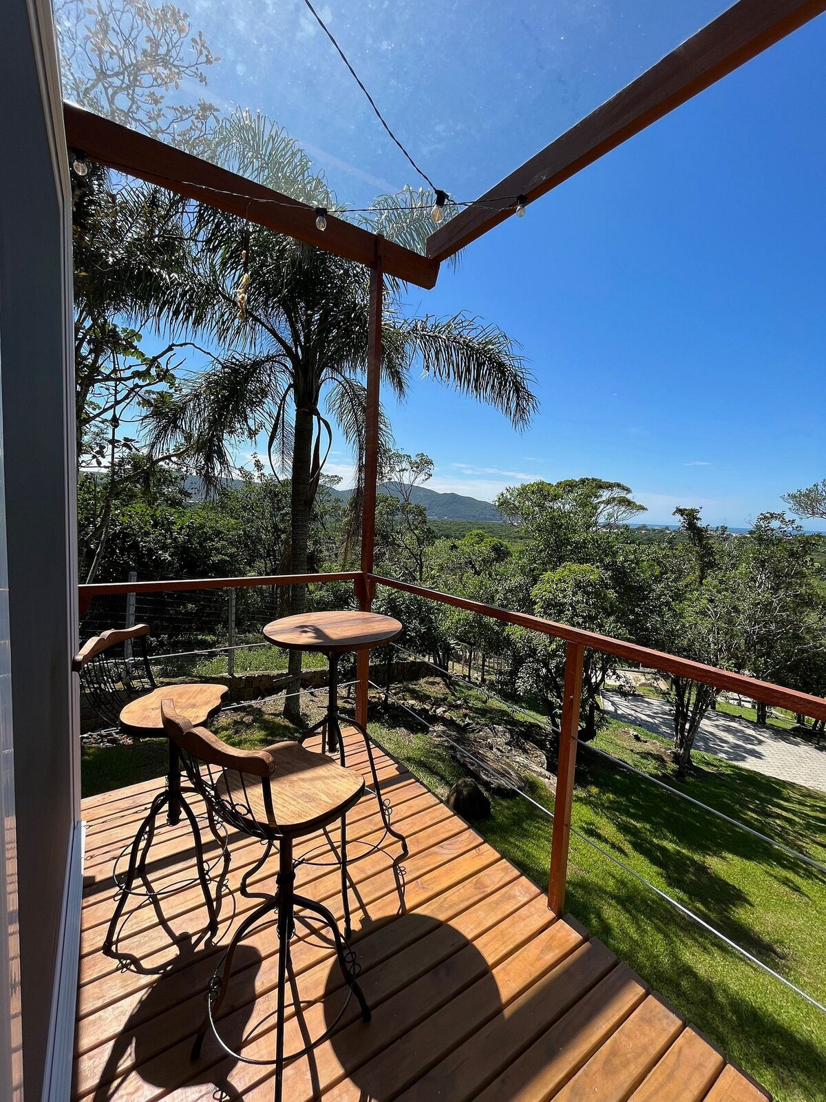 A balcony area is seen with two bar stools positioned at a wooden table. Sunlight floods the space, offering views of lush greenery and distant hills, framed by palm trees and a clear blue sky.