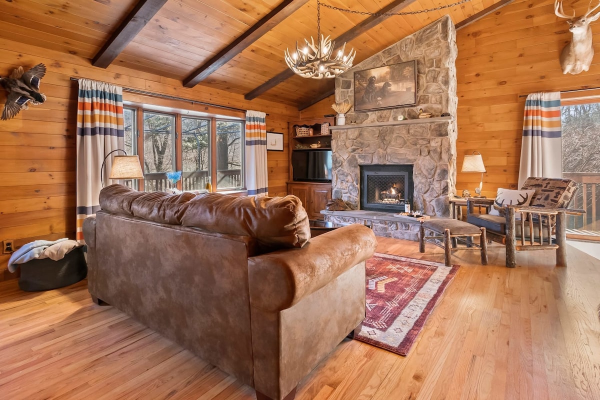 A rustic living area features a stone fireplace, enhancing the cabin's cozy ambiance. Large windows allow natural light to fill the space. A comfortable leather sofa provides seating, accompanied by wooden chairs and a warm area rug. Decorative wildlife mounts add character to the walls.