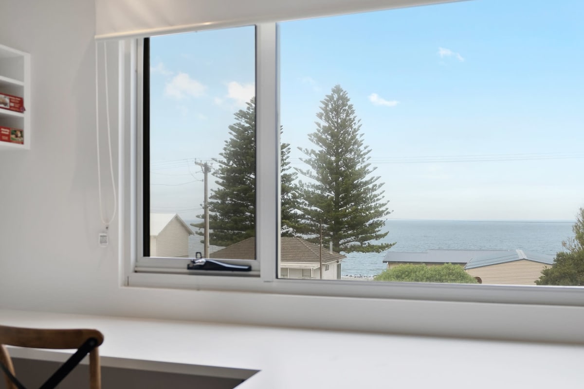 A view from a desk area showcases a large window overlooking the ocean. Tall pine trees are visible against a clear blue sky, adding a natural touch to the serene water view. The workspace features a minimalist design with a simple wooden chair.