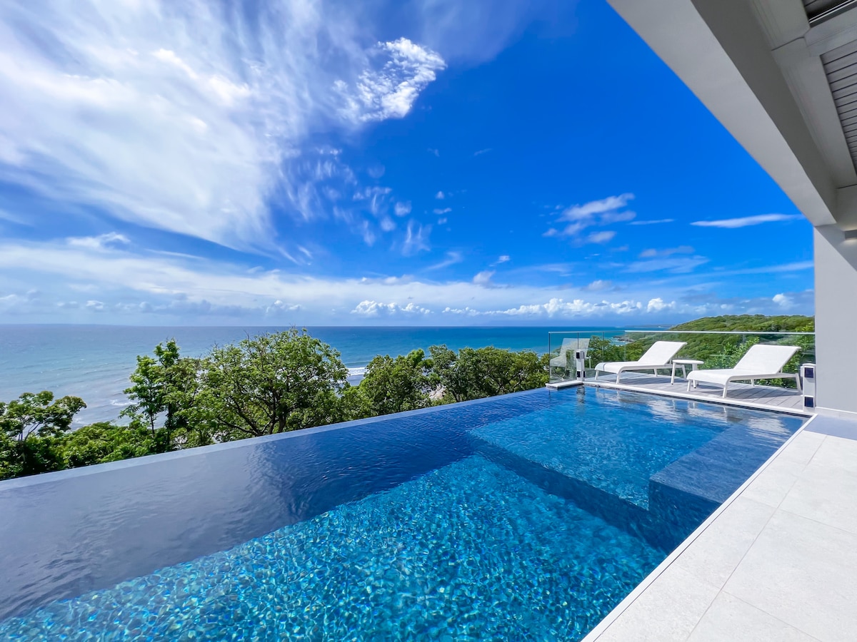 An expansive infinity pool reflects the clear blue sky and offers a panoramic view of the ocean. Sun loungers are positioned beside the water, inviting relaxation. Lush greenery surrounds the area, enhancing the tranquil atmosphere.