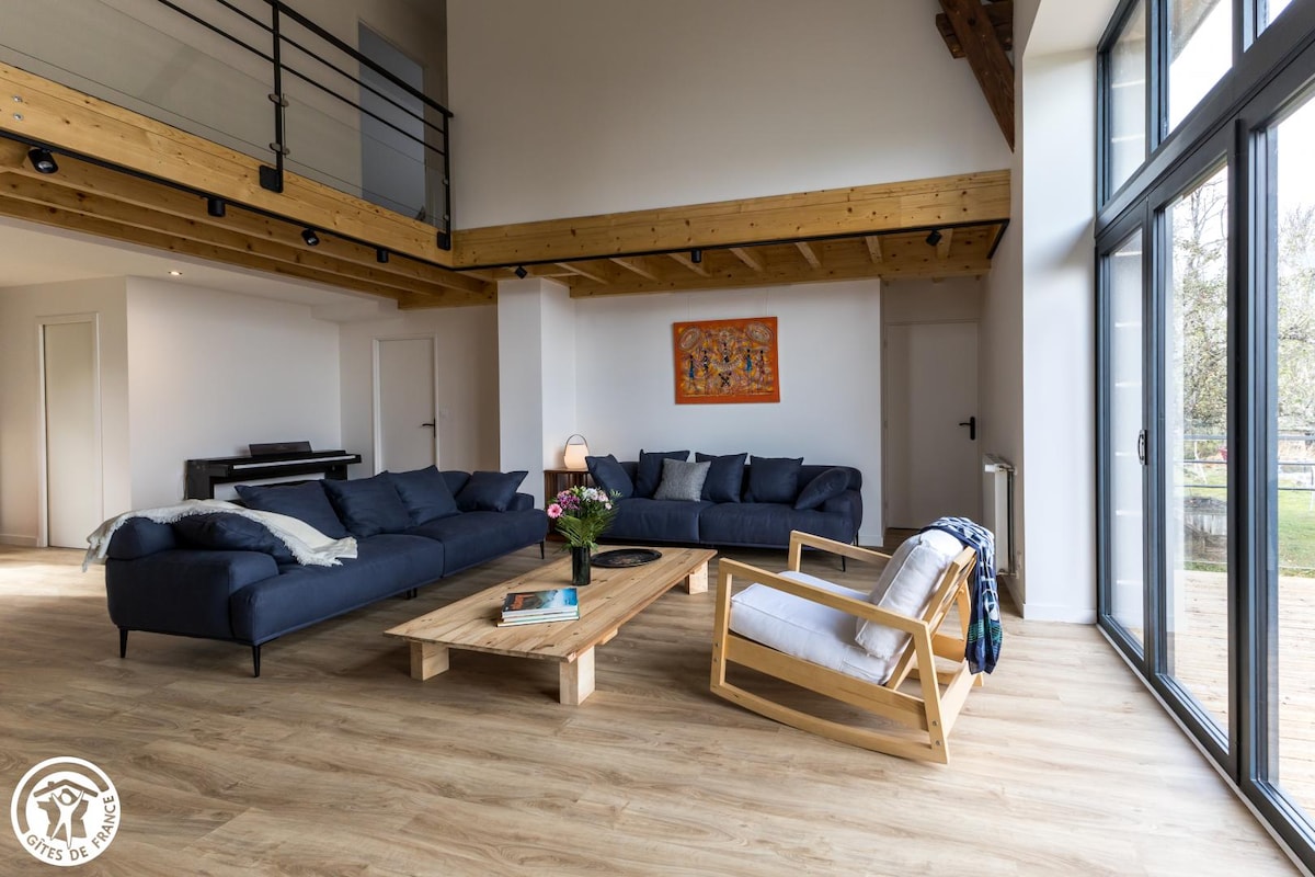 A spacious living area features a large blue sectional sofa arranged around a wooden coffee table. Natural light streams through expansive glass doors, illuminating the light-colored wooden flooring. A comfortable armchair adds to the inviting atmosphere, while artwork adorns the wall in the background.
