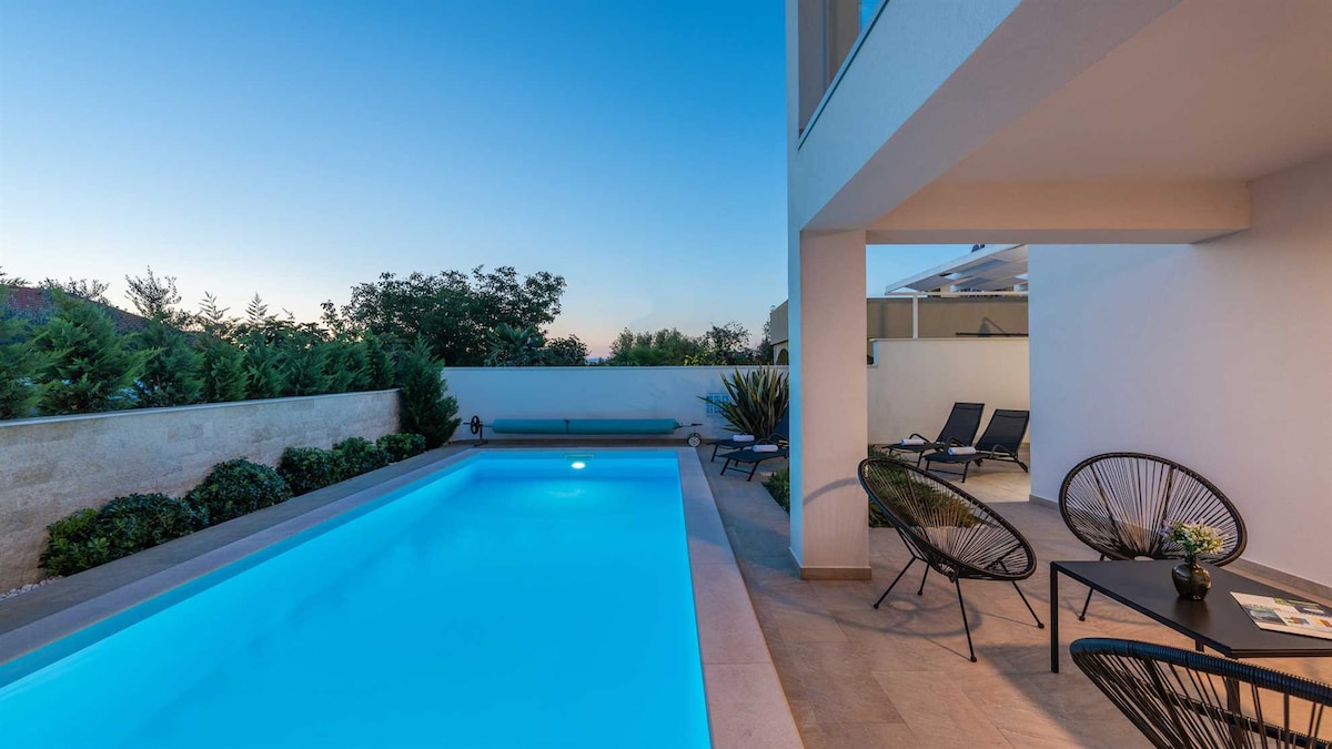 A private pool is nestled beside a clean, modern villa, surrounded by greenery. Comfortable lounge chairs are positioned for relaxation, and a small table with decorative items invites guests to enjoy the outdoor space. The area is softly illuminated in blue as evening approaches.