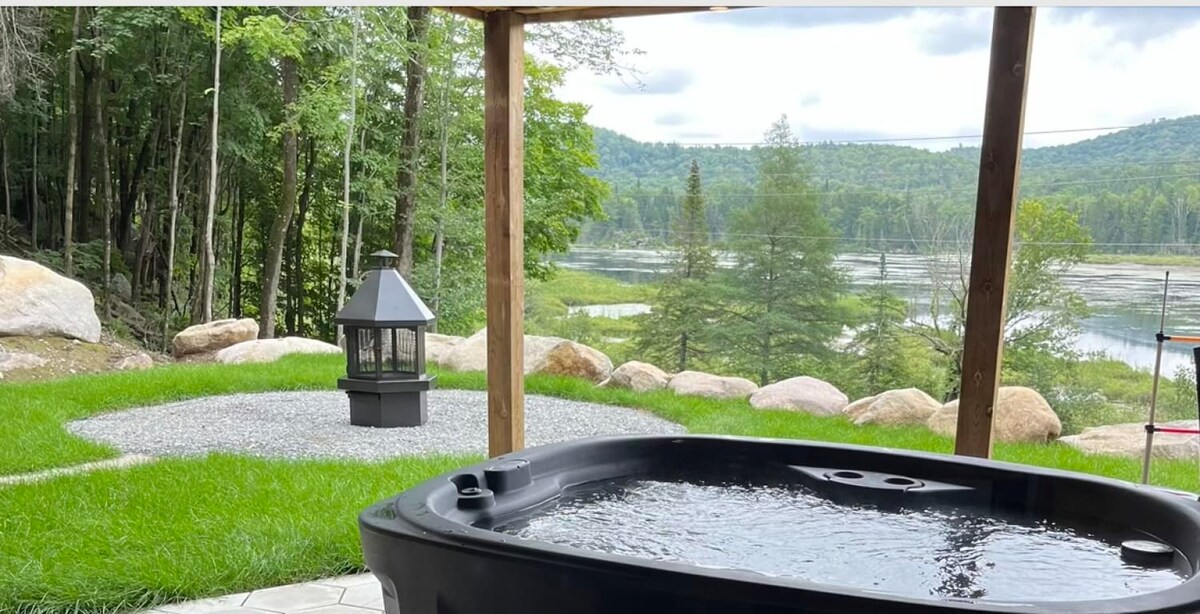 A private hot tub is positioned beneath a wooden structure, featuring calm water that reflects the surrounding greenery. In the background, a lantern sits beside an outdoor fire area, with a view of a serene landscape and trees lining a body of water.