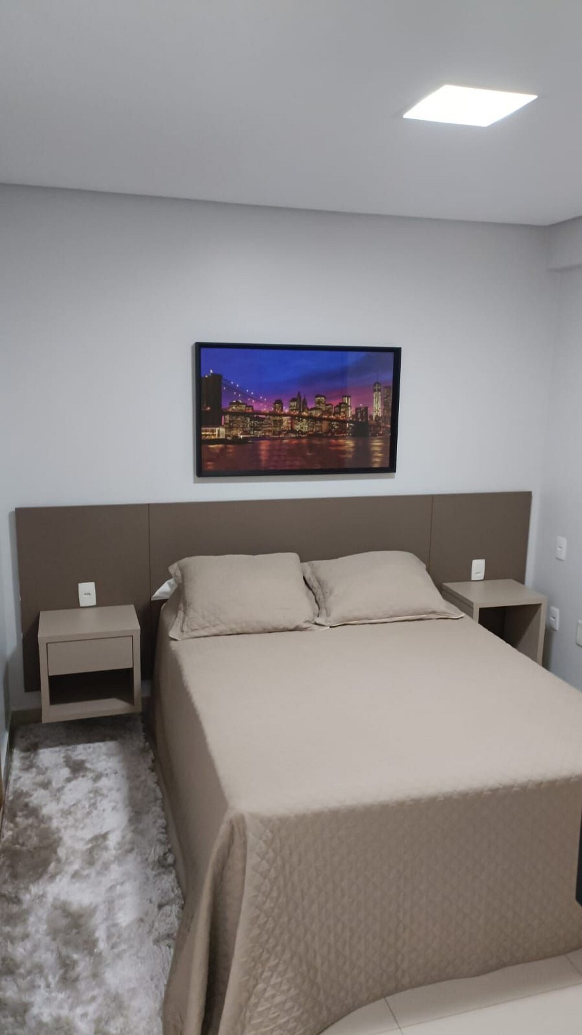 A cozy bedroom is presented with a spacious bed covered with a neutral-toned blanket. Nightstands flanking the bed house convenient bedside lamps. A colorful cityscape artwork adorns the wall above the bed, complementing the light gray color of the surrounding walls.