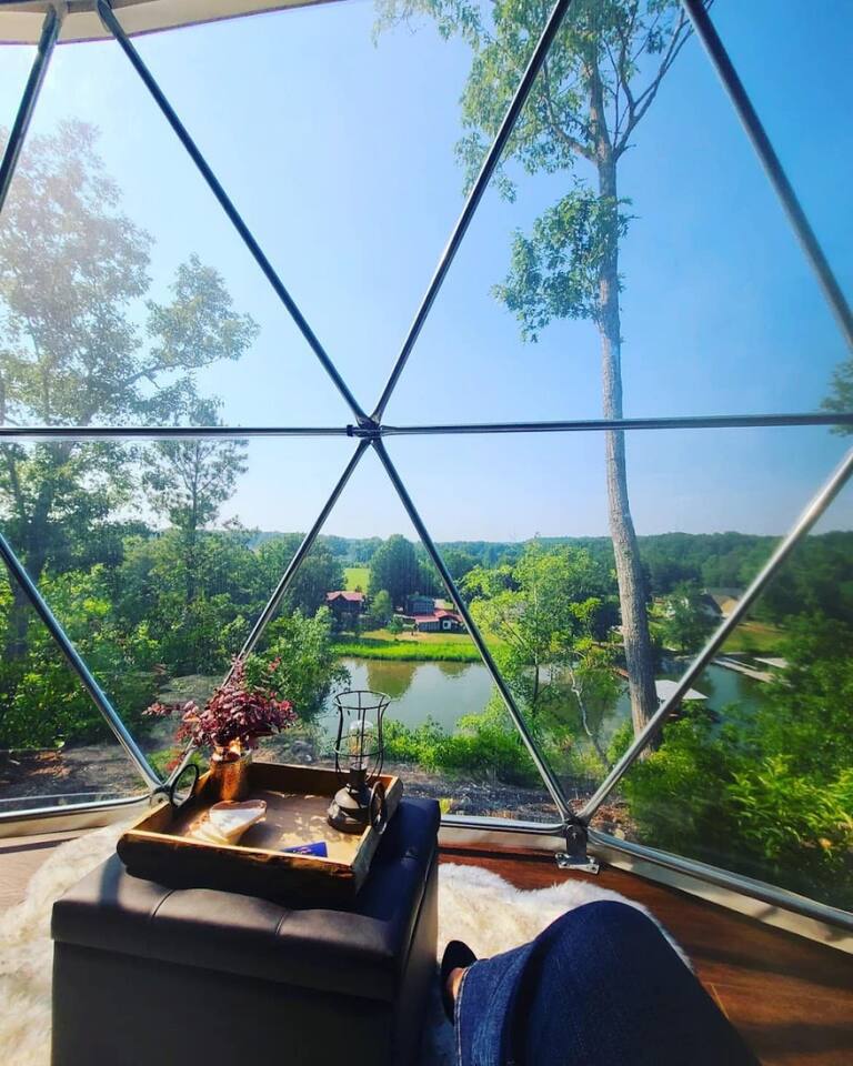 A clear view through large triangular windows showcases lush greenery and a serene waterway. A small table with a decorative vase and candle is positioned in the foreground, inviting relaxation while enjoying the scenic landscape.
