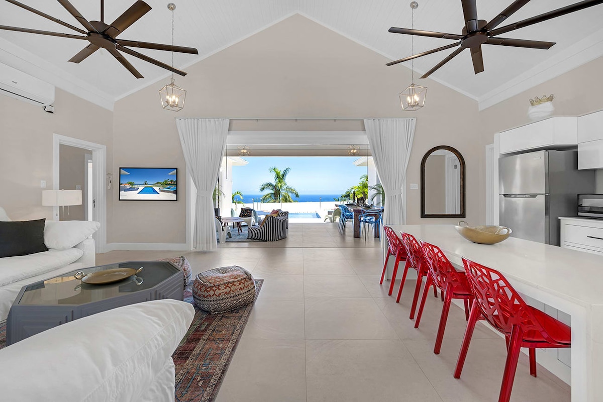An open-concept living space is displayed with a view of the Caribbean Sea beyond large glass doors. Modern furniture includes a sectional white sofa, a glass coffee table, and a dining area with red chairs. Ceiling fans are mounted above, providing a breezy atmosphere.