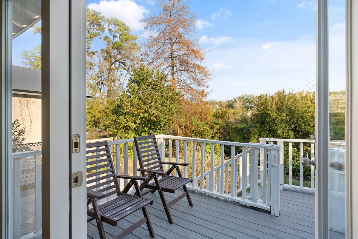 A private balcony is visible, featuring two wooden lounge chairs that face a lush greenery backdrop. The deck overlooks a garden area with trees, and a clear blue sky brings natural light to the space.