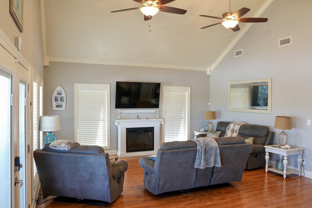A spacious living area features two cozy sofas positioned around a coffee table, with a large flat-screen television mounted on a light gray wall. Natural light enters through two windows, and a decorative mirror adds depth. A ceiling fan enhances airflow.
