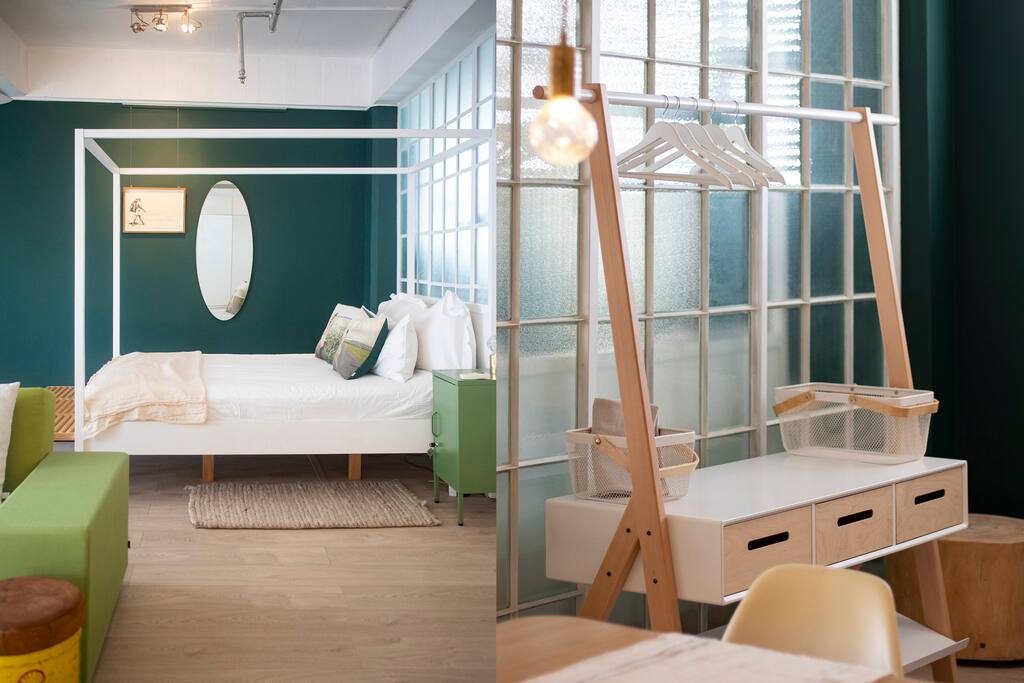 A modern sleeping area features a four-poster bed with crisp white linens, accompanied by soft green accents. A compact desk with storage options stands nearby, along with a hanging rod for clothes. Light filters through large glass windows, creating an airy atmosphere.