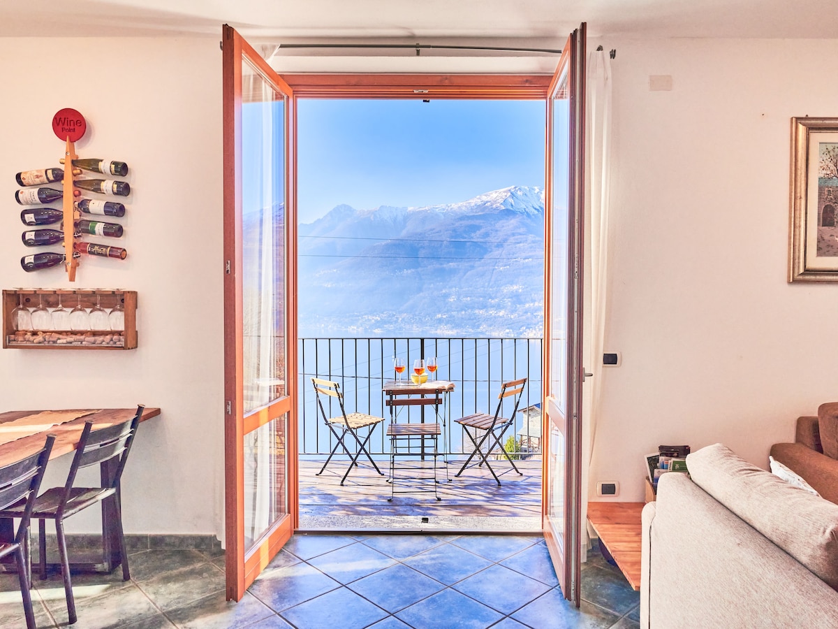Glass doors open to reveal a scenic balcony view, featuring a table set with two drinks and chairs. The surrounding mountains and lake can be seen in the distance, enhancing the tranquil atmosphere of the living space.