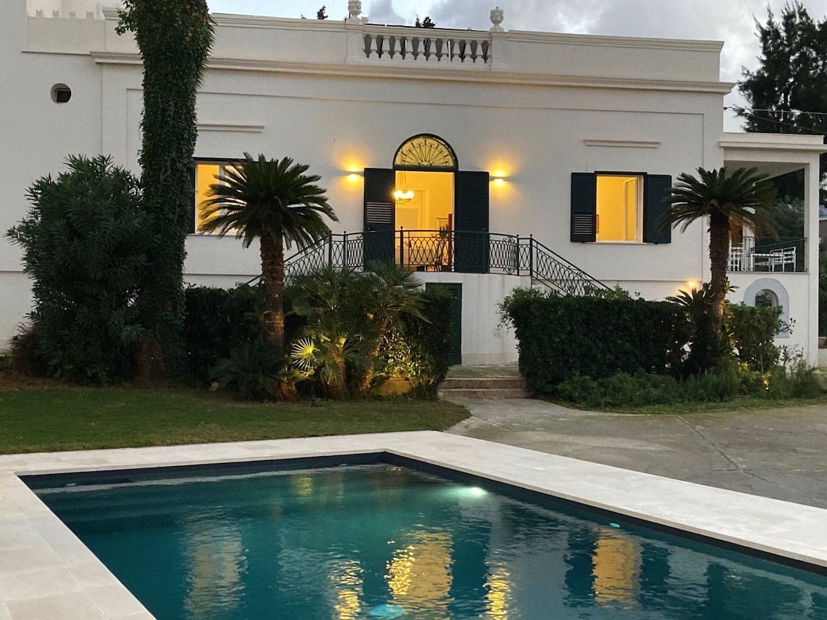The exterior of the villa is depicted, showcasing a classic structure with large windows and an elegant balcony. A private swimming pool is in the foreground, surrounded by lush greenery. Warm light emanates from the villa's windows, creating a welcoming ambiance.