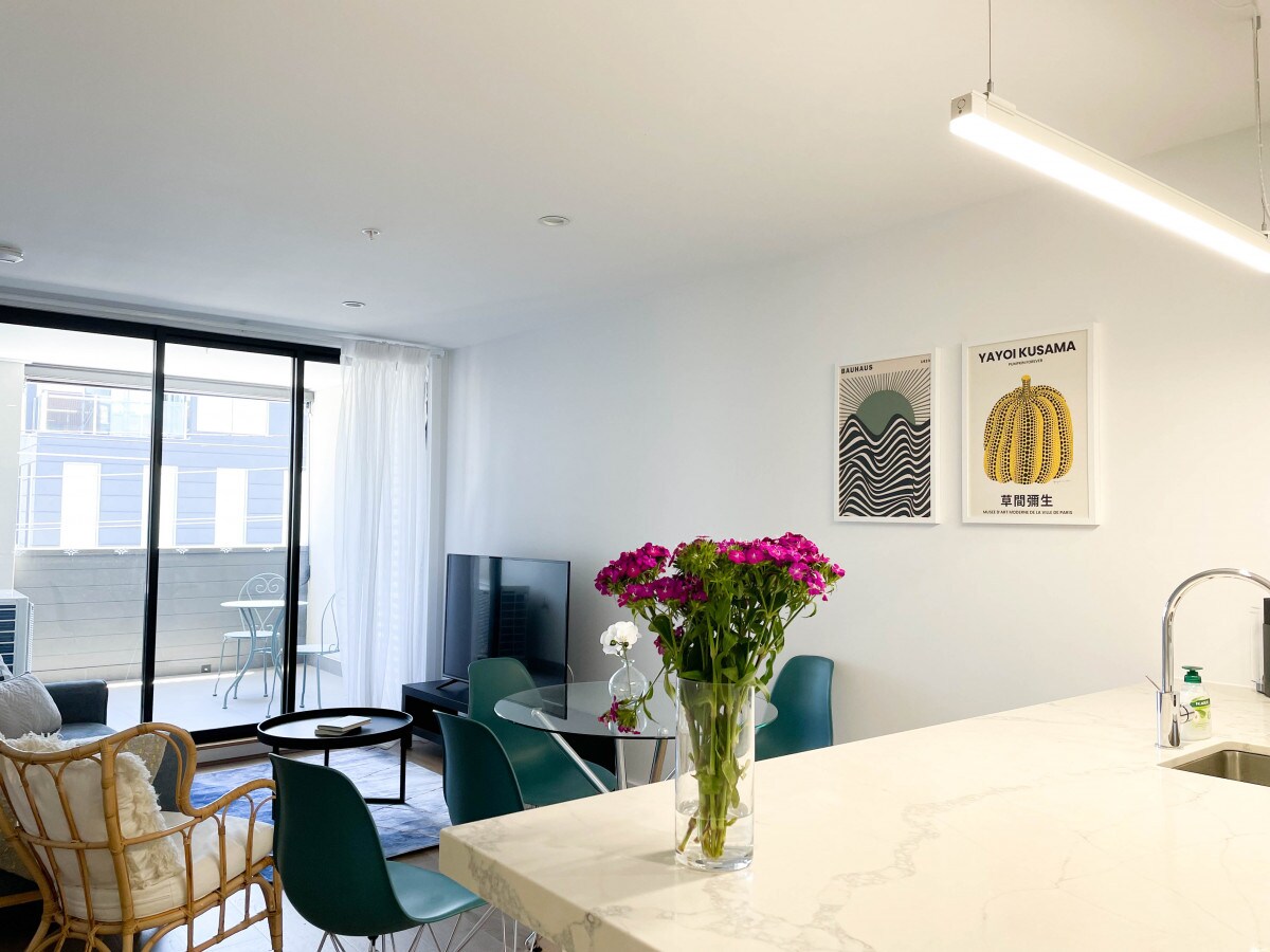 A modern living space showcases a marble countertop with a stylish vase of flowers. In the background, a glass sliding door opens to a balcony area with outdoor seating. Artwork adorns the walls, enhancing the contemporary atmosphere.