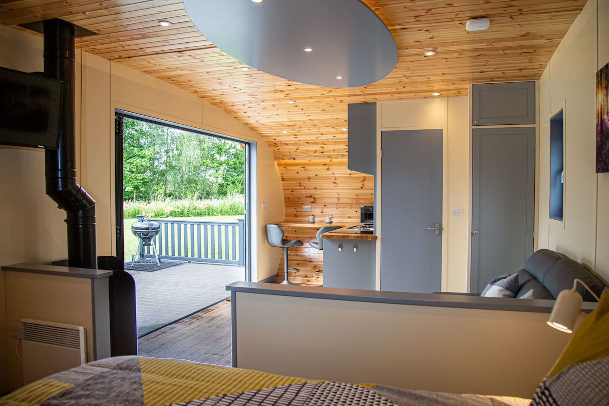 The interior features wooden paneling with a stylish curved ceiling. A spacious bed is visible, with a sitting area and TV nearby. Bifold doors open to a decked area with outdoor seating and a barbecue. A small kitchenette is positioned to the side.
