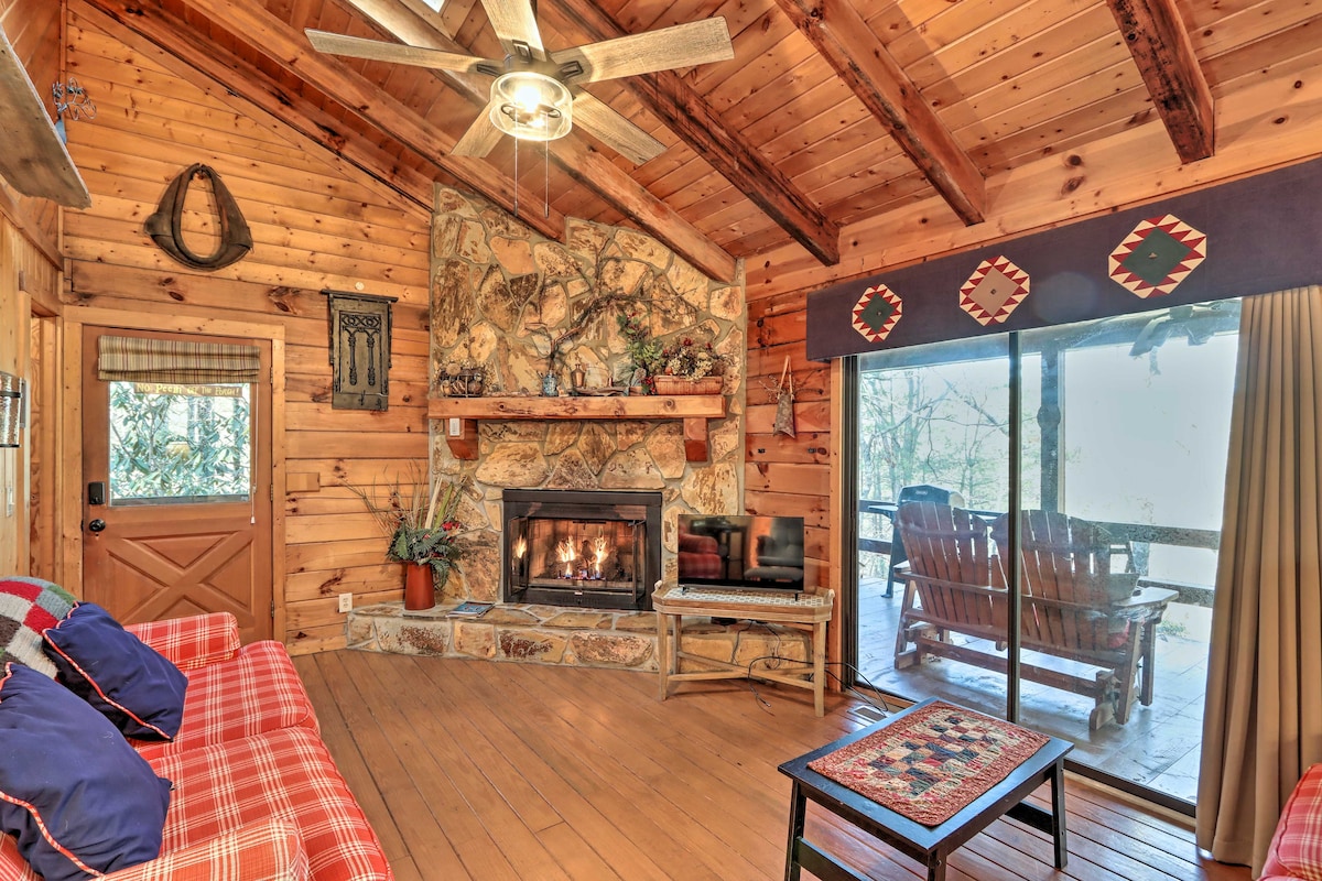 The living area showcases a stone fireplace, flanked by a comfortable seating arrangement with plaid and solid cushions. A ceiling fan hangs above, and large glass doors provide natural light while offering access to an outdoor space featuring wooden rocking chairs.