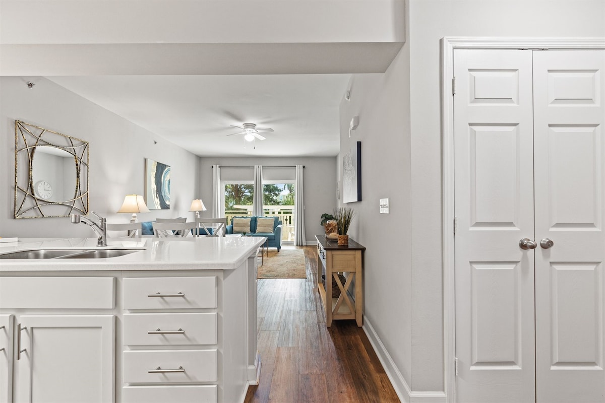 The open concept living space is framed by a white kitchen with modern cabinetry and a contrasting countertop. A cozy seating area is decorated in soft blue tones, with large windows allowing natural light to illuminate the room.