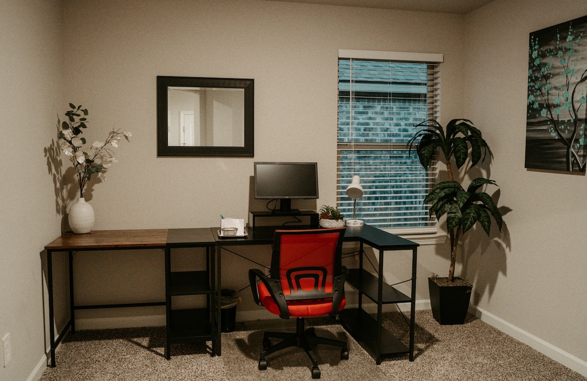 A dedicated workspace features a modern desk setup with a black chair accented in red. A computer monitor and lamp are positioned on the desk, alongside decorative plants. Natural light filters through the window, complemented by soft-colored walls.