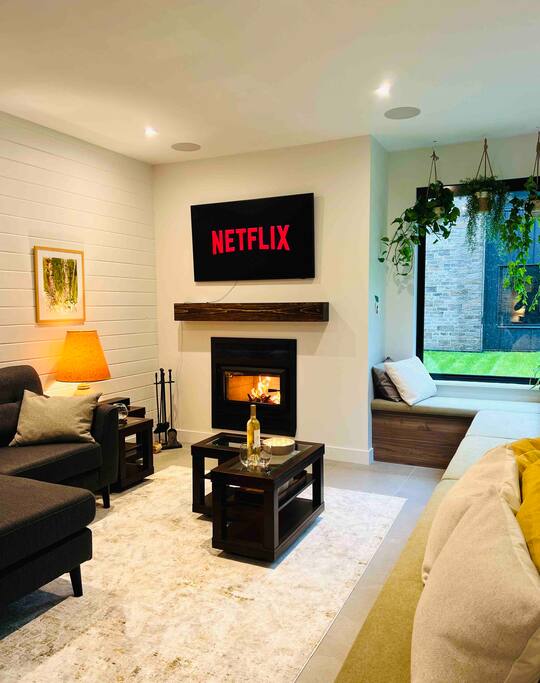 A modern living area features a flat-screen television displaying the Netflix logo above a wood-burning fireplace. Comfortable seating includes a dark sofa and light-colored armchair, complemented by a small coffee table. Natural light is welcomed through a large window, enhancing the inviting space.