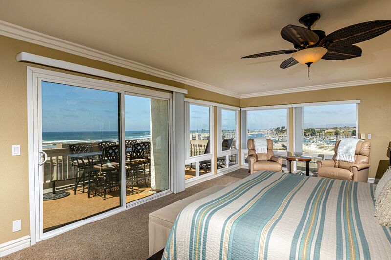 The primary suite features a spacious bed with light-colored bedding. Large windows provide panoramic ocean views, while a sliding door leads to a balcony with outdoor seating. Two recliners are positioned next to the windows, allowing for relaxation and enjoying the scenery.