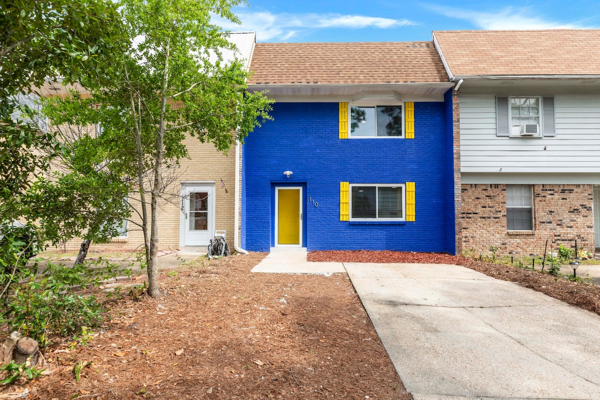 A vibrant blue exterior is complemented by bright yellow shutters and a cheerful front door. The property features a well-maintained driveway leading up to the entrance, with landscaping that includes mulch and greenery on either side.