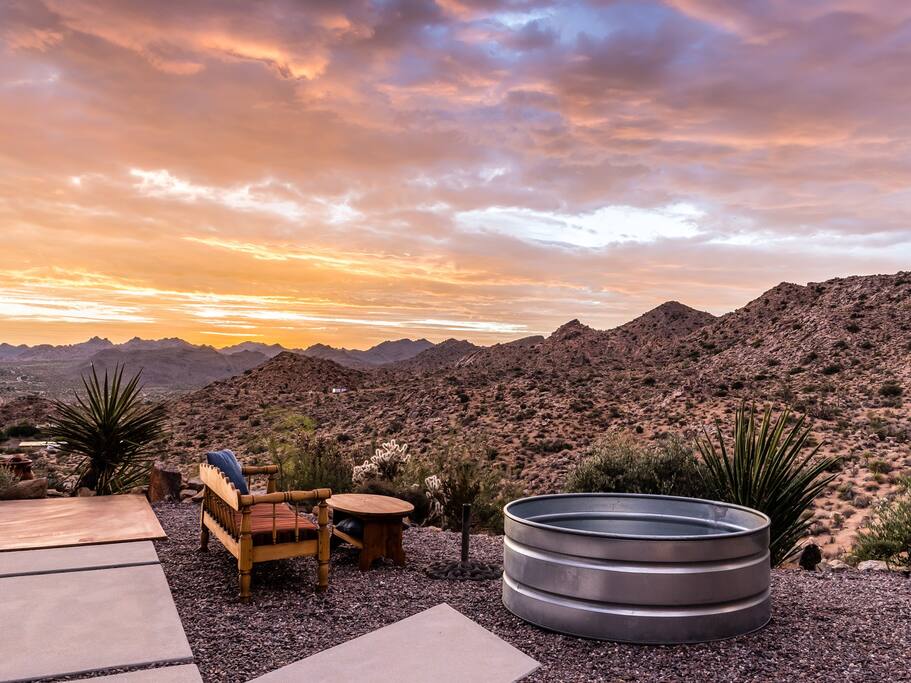 A cowboy tub is positioned beside a small seating area, both framed by desert vegetation. The backdrop showcases rolling mountains under a vibrant sunset, with clouds reflecting shades of orange and purple across the sky.