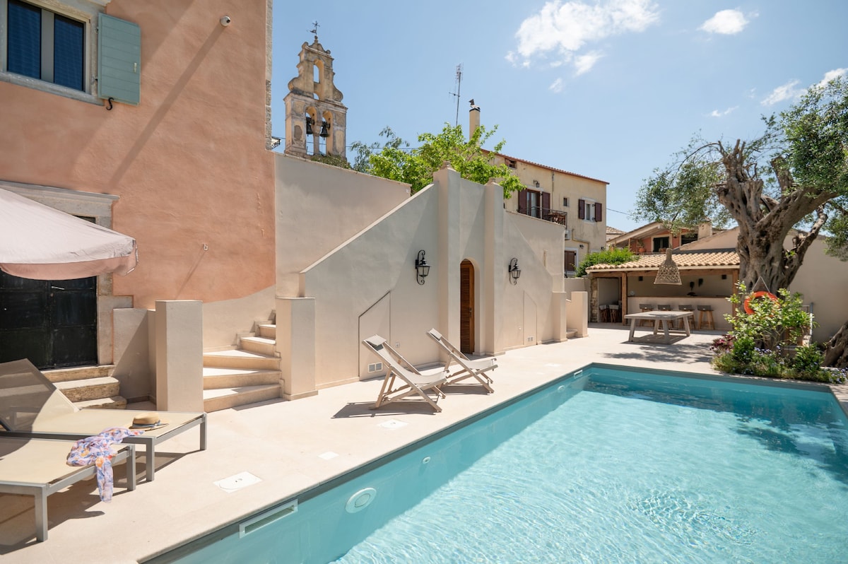 The private heated pool is surrounded by a spacious deck, featuring sun loungers and a dining area. Soft sunlight reflects off the clear water, while a nearby staircase leads to a charming terrace. A historic building and trees can be seen in the background.