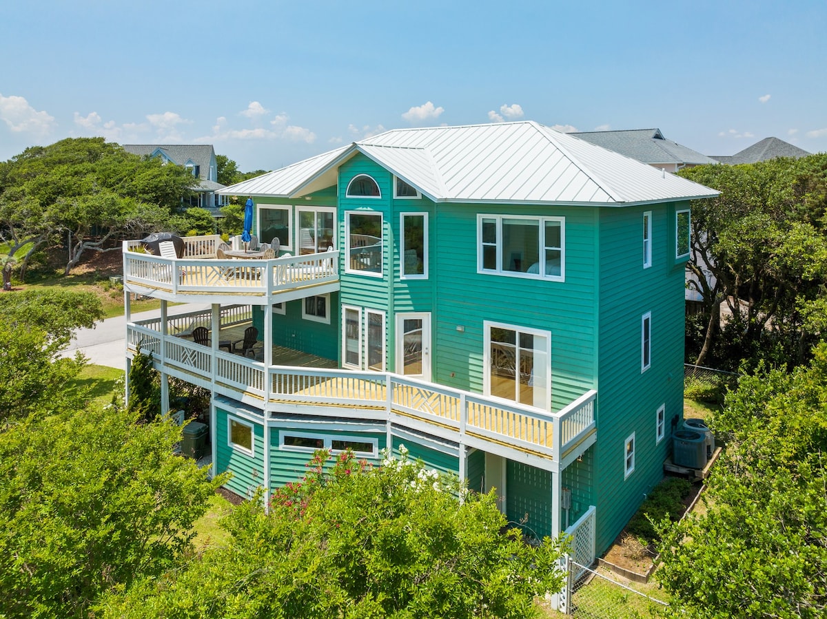 A spacious oceanview home in a vibrant teal exterior is shown from an angle highlighting its three levels. Multiple open and covered decks are visible, adorned with outdoor furniture. Surrounding greenery completes the aesthetic of this coastal retreat.