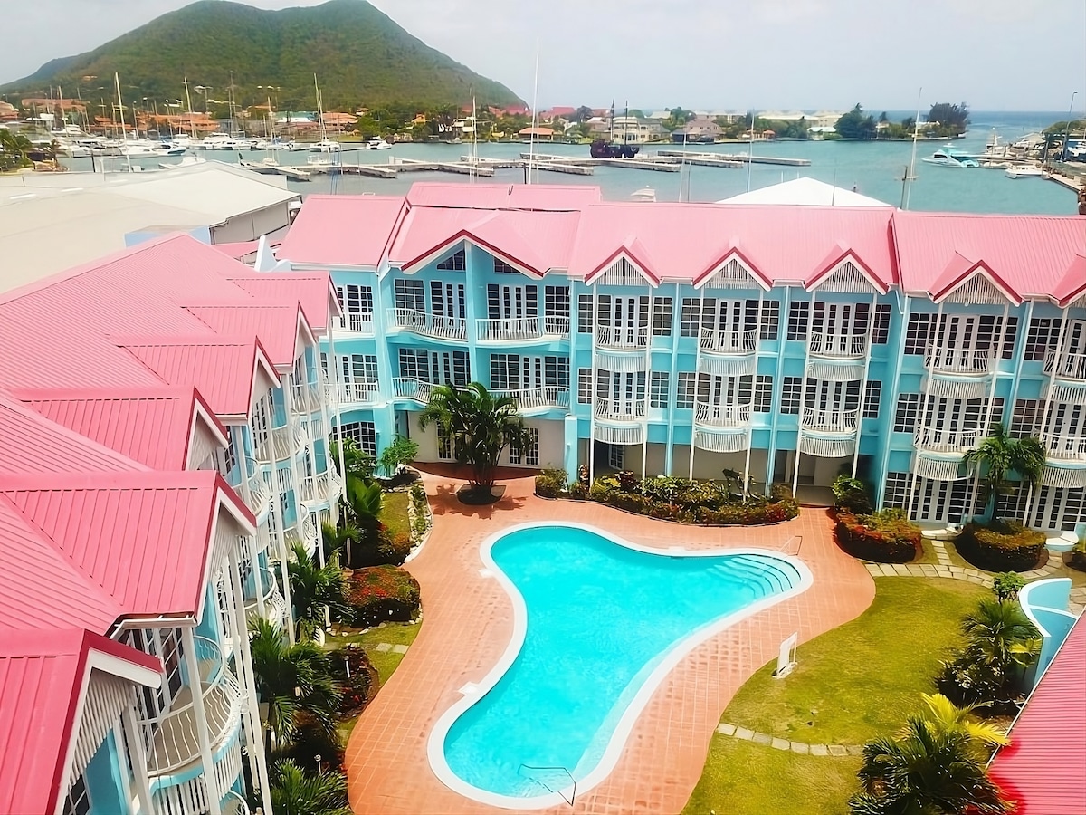 An aerial view captures a vibrant hotel complex with light blue and pink accents. The outdoor swimming pool is framed by a lush garden, with palm trees and flowering plants. In the background, the marina and hills create a scenic coastal backdrop.