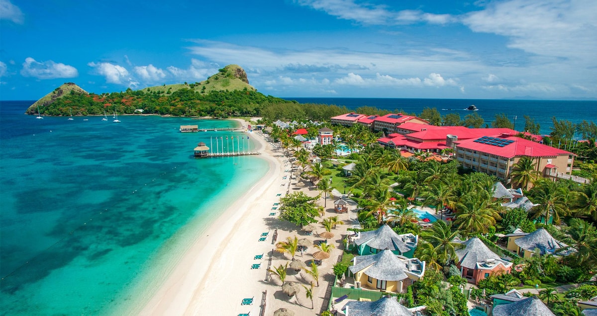 An aerial view captures the resort's beachfront, showcasing soft sandy shores and vibrant turquoise waters. Lush palm trees frame the landscape, while the resort buildings feature a striking red roof, blending harmoniously with the surrounding natural beauty. Nearby hills create a scenic backdrop.