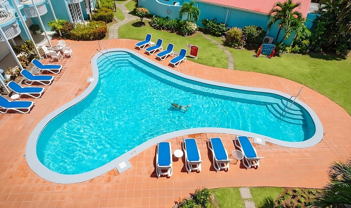 A spacious swimming pool in a unique shape is surrounded by sun loungers with blue cushions. Lush greenery is visible around the pool area, enhancing the outdoor relaxation space. The clear water of the pool reflects the bright sky above.