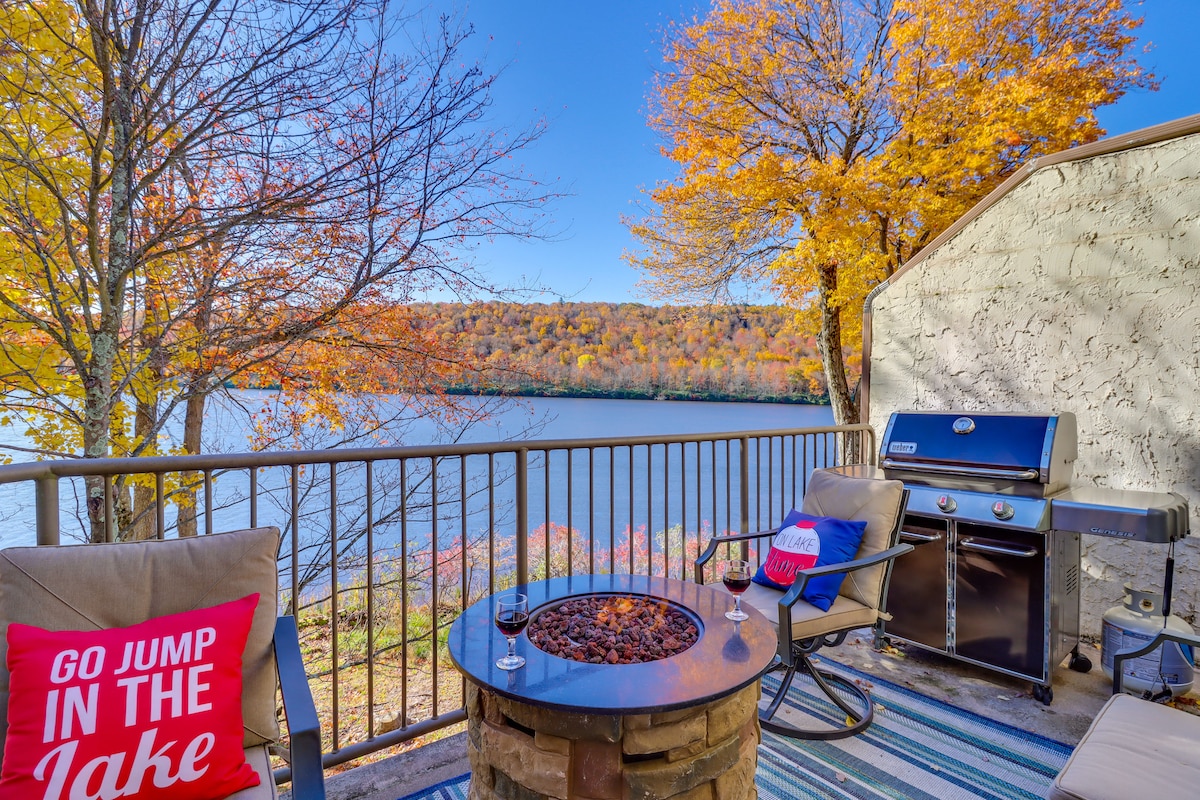 A lakeside patio features two comfortable chairs beside a fire table, with a grill nearby. Colorful autumn foliage frames the scenic view of the lake, reflecting the vibrant fall colors. Soft rugs add texture to the outdoor space, inviting relaxation.
