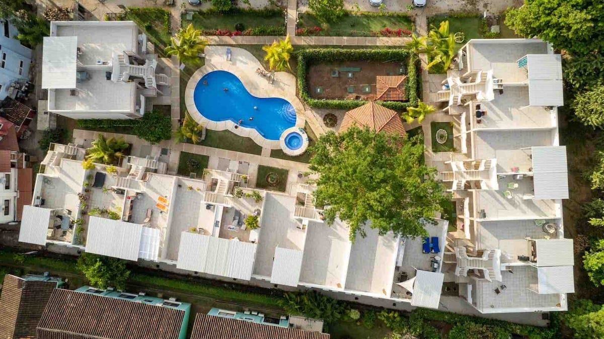 An aerial view of the Flor de Limon complex showcases the layout of the two-story condos surrounding a central pool area. Lush greenery decorates the grounds, with palm trees and garden areas creating a tranquil setting. The inviting blue pool is centrally located, offering relaxation options.