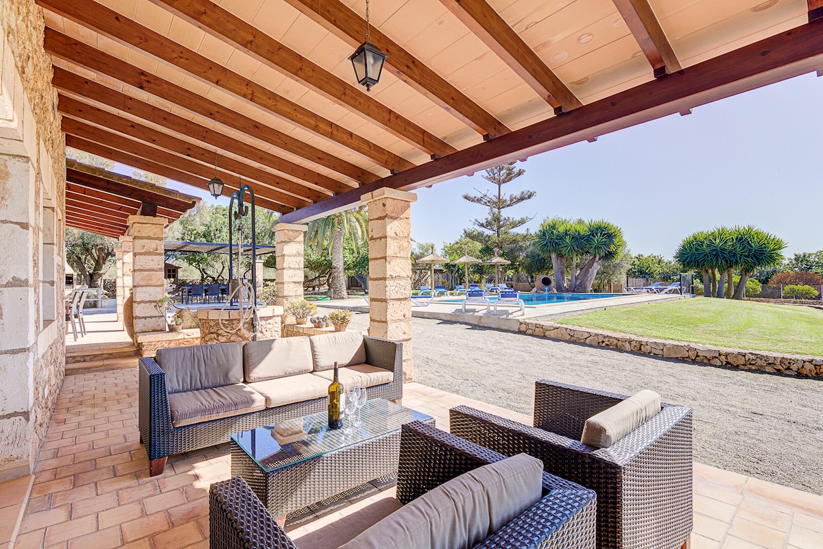 A shaded outdoor terrace is furnished with comfortable seating arranged around a glass-topped table. The space offers views of the well-maintained garden and swimming pool area, with several sun loungers and landscaped greenery visible in the background.