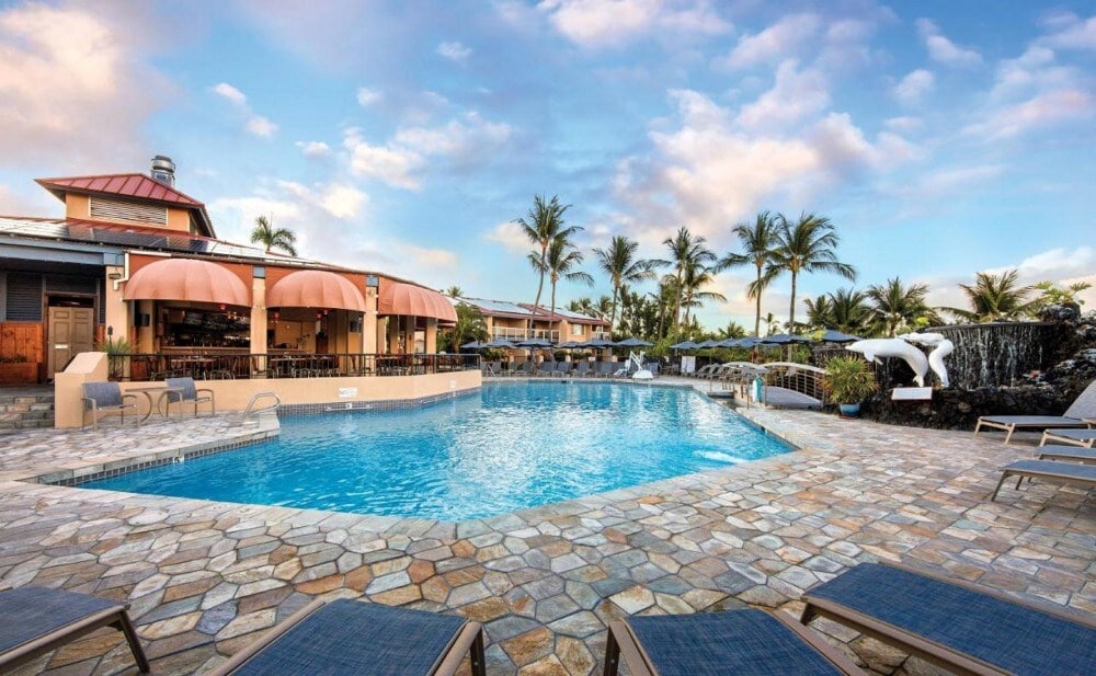 A shared outdoor pool is surrounded by natural stone paving and lounge chairs. Tropical palm trees are visible in the background, and a restaurant with arched awnings overlooks the pool area, providing a relaxing environment for guests.