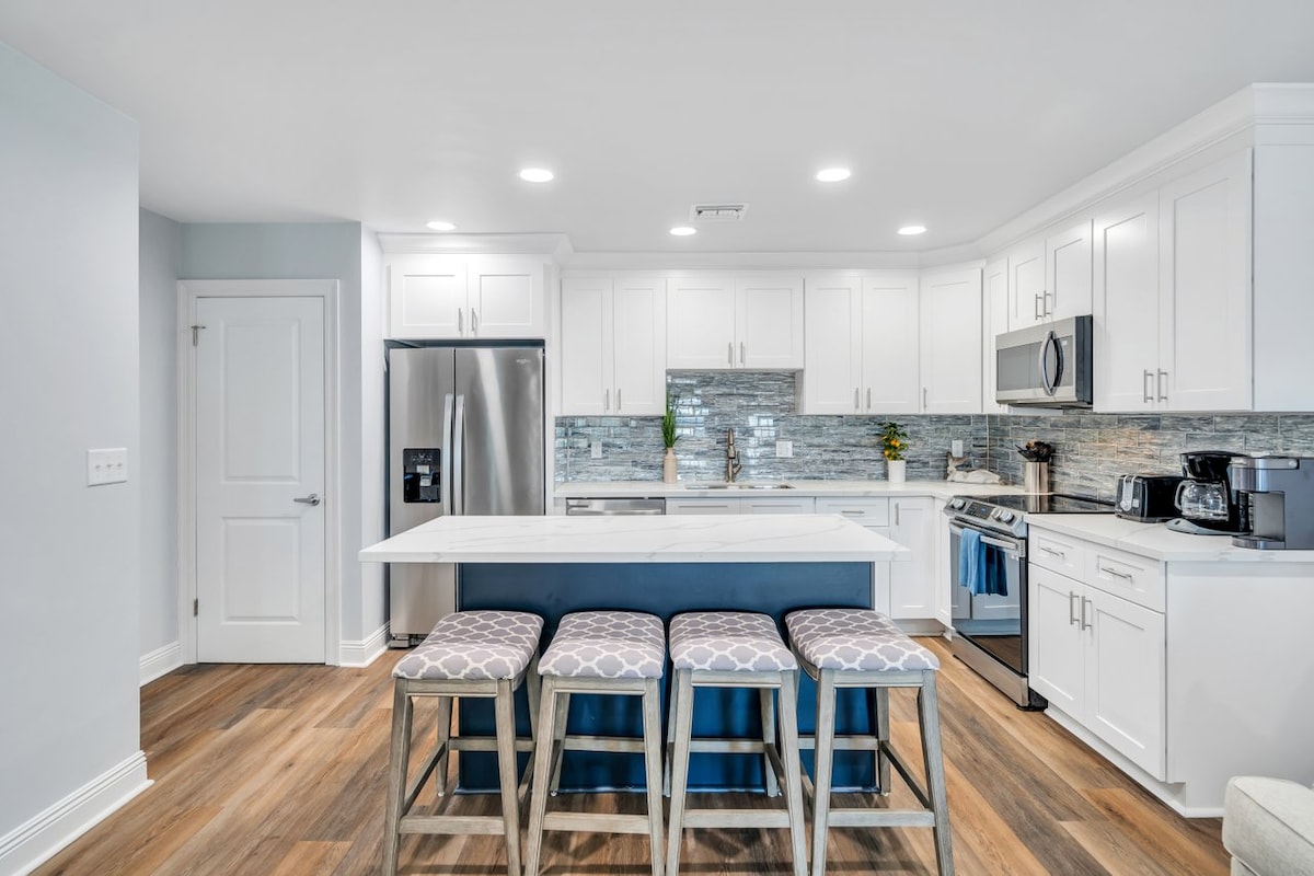 A modern kitchen features sleek white cabinetry and stainless-steel appliances. A large island with seating for four is positioned center stage. The backsplash consists of gray and white tiles, seamlessly complementing the wood flooring throughout the space.