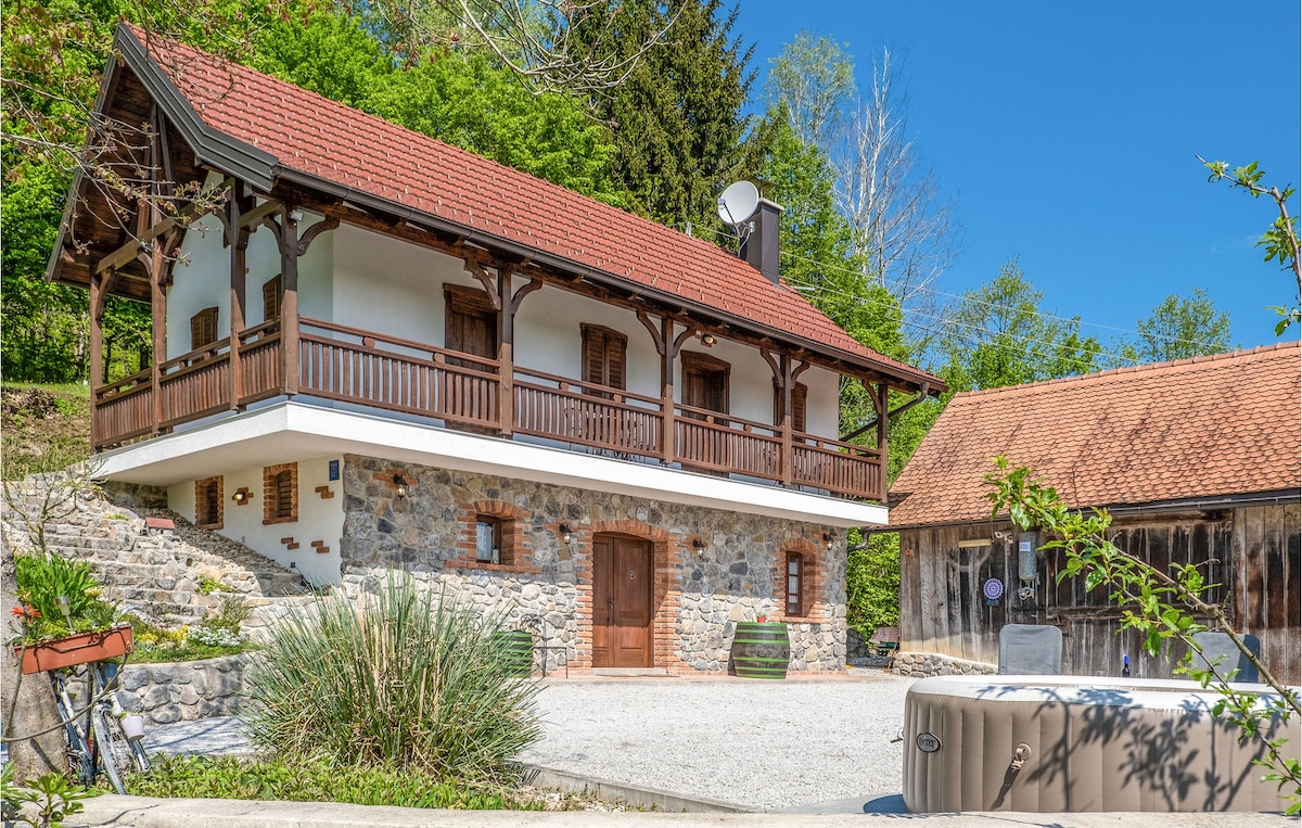 A charming cottage is featured with a mix of stone and wooden elements. The two-story structure is complemented by a rustic barn nearby. A well-maintained garden surrounds the home, with a jacuzzi visible in the foreground, set against a backdrop of lush greenery and a clear blue sky.