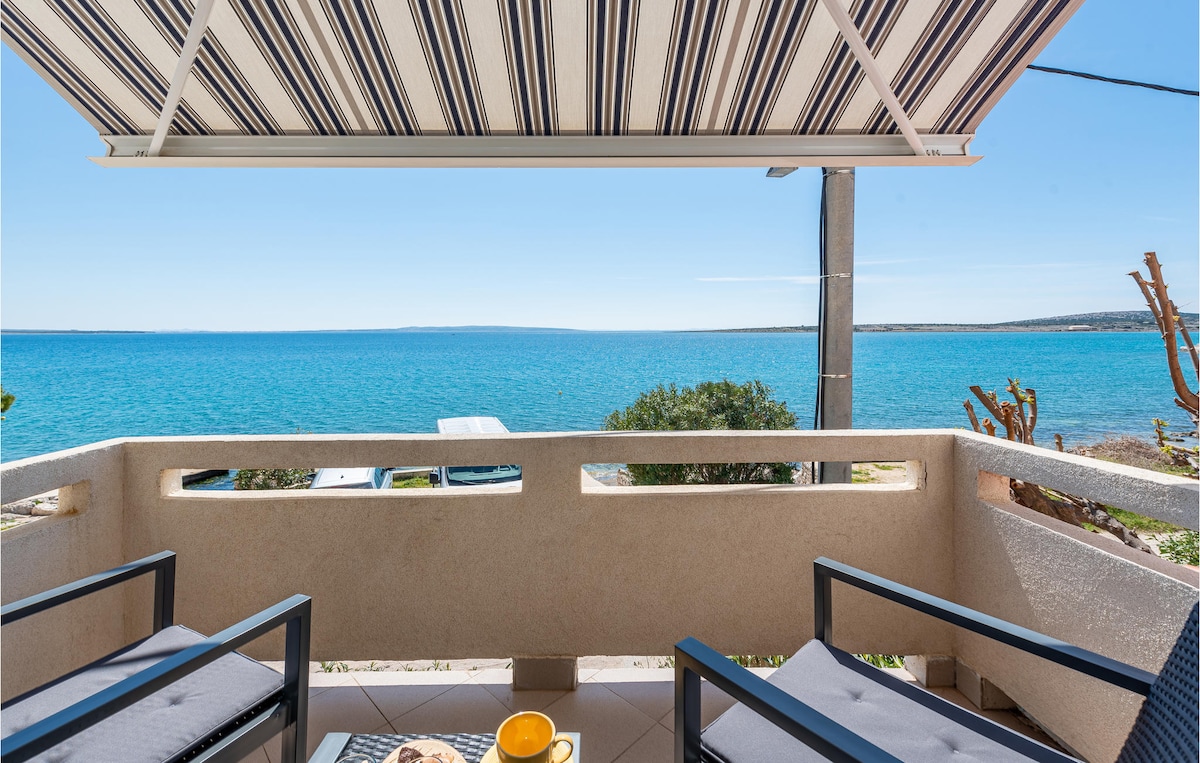 An outdoor terrace features two comfortable chairs and a small table. The awning provides shade, while a stunning sea view stretches across the horizon. Potted plants and a partial view of the coastline enhance the inviting setting.