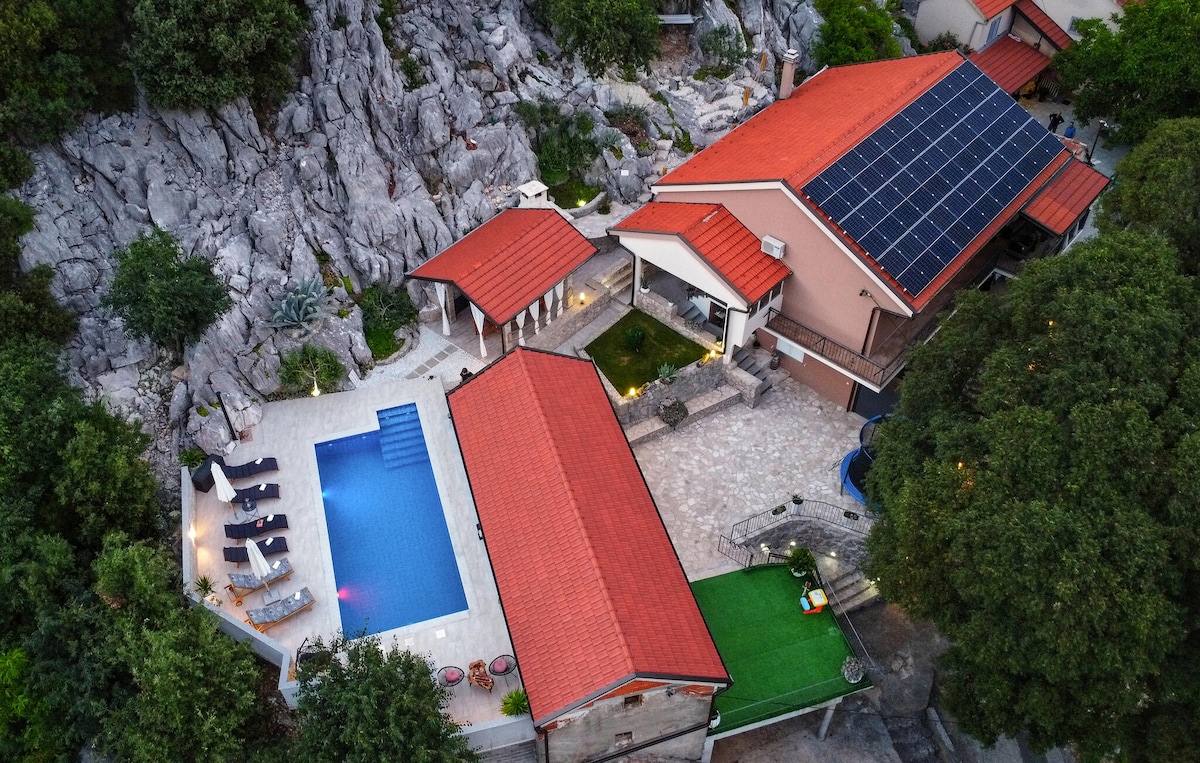An aerial view captures a modern holiday home nestled among rocky terrain. The red-tiled roofs contrast with the greenery. A spacious pool is visible, flanked by loungers, while a summer kitchen area and a garden can also be seen in the layout.