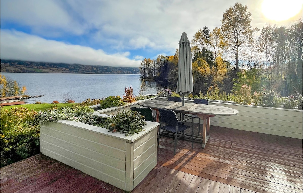 A spacious terrace overlooks the tranquil lake, featuring a large dining table surrounded by comfortable chairs. An umbrella provides shade, while lush greenery and autumn trees frame the scene, creating a peaceful atmosphere for outdoor meals and relaxation.