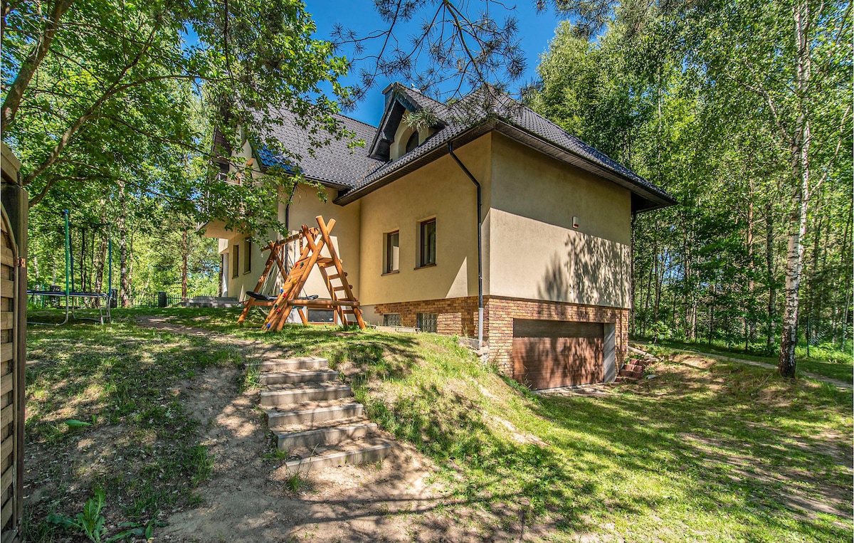 A spacious cottage is set among lush greenery, featuring a two-story structure with a sloped black roof. Steps lead from the foreground to the entrance, where a wooden climbing frame and slide are visible, suggesting a family-friendly environment.