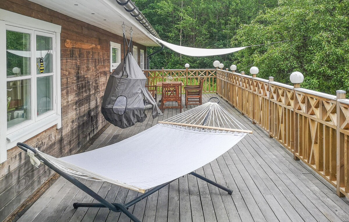 A spacious wooden terrace features a comfortably suspended hammock and a hanging chair, surrounded by a wooden railing. A dining set is visible in the background, with spherical lights adding subtle illumination. Lush greenery provides a serene backdrop, enhancing the outdoor space.