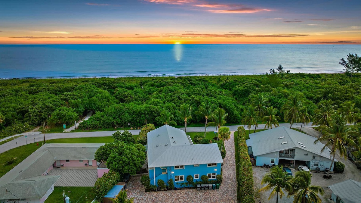 An aerial view captures the Surfside Beach House in the foreground, nestled among lush greenery, with a stunning ocean horizon painted in warm orange and cool blue hues at sunset. The clear sky complements the peaceful coastal setting, showcasing the nearby beach and palm trees.