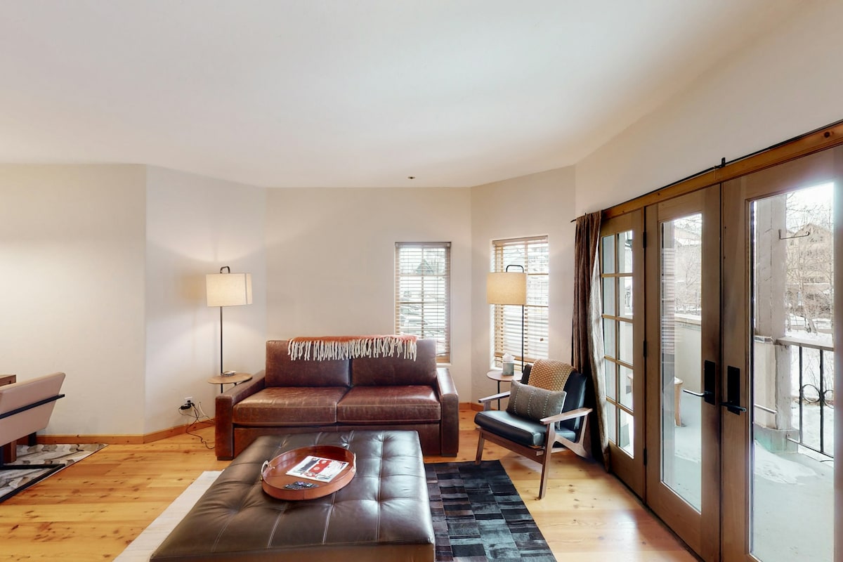 A living room area is presented, featuring a brown leather sofa and a wooden coffee table with a decorative tray. Natural light enters through the glass doors, which lead to a balcony. A side chair and a floor lamp enhance the cozy atmosphere.