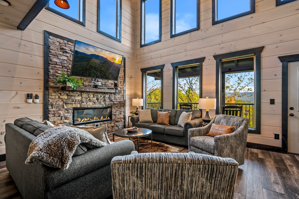 A spacious living area is adorned with large windows, offering scenic views of the mountains. A stone fireplace provides warmth, complemented by a wall-mounted large television. Comfortable seating includes a plush sectional sofa and two accent chairs, inviting relaxation in a cozy atmosphere.