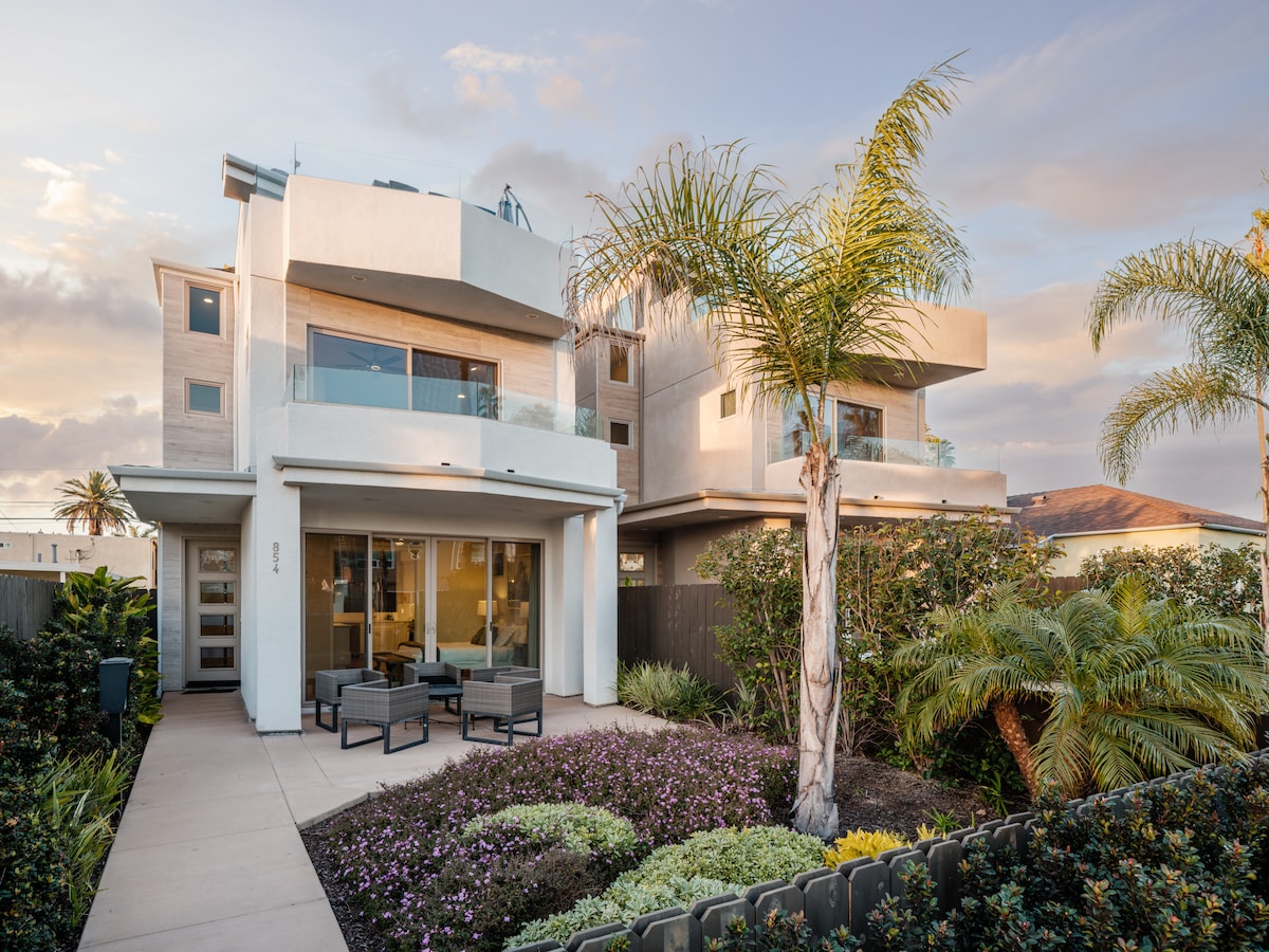 The exterior of a modern three-story home is presented, surrounded by lush greenery and colorful plants. A patio area with comfortable seating is visible, offering a spot to enjoy the landscaped yard. Large windows reveal an open, airy design, inviting natural light.