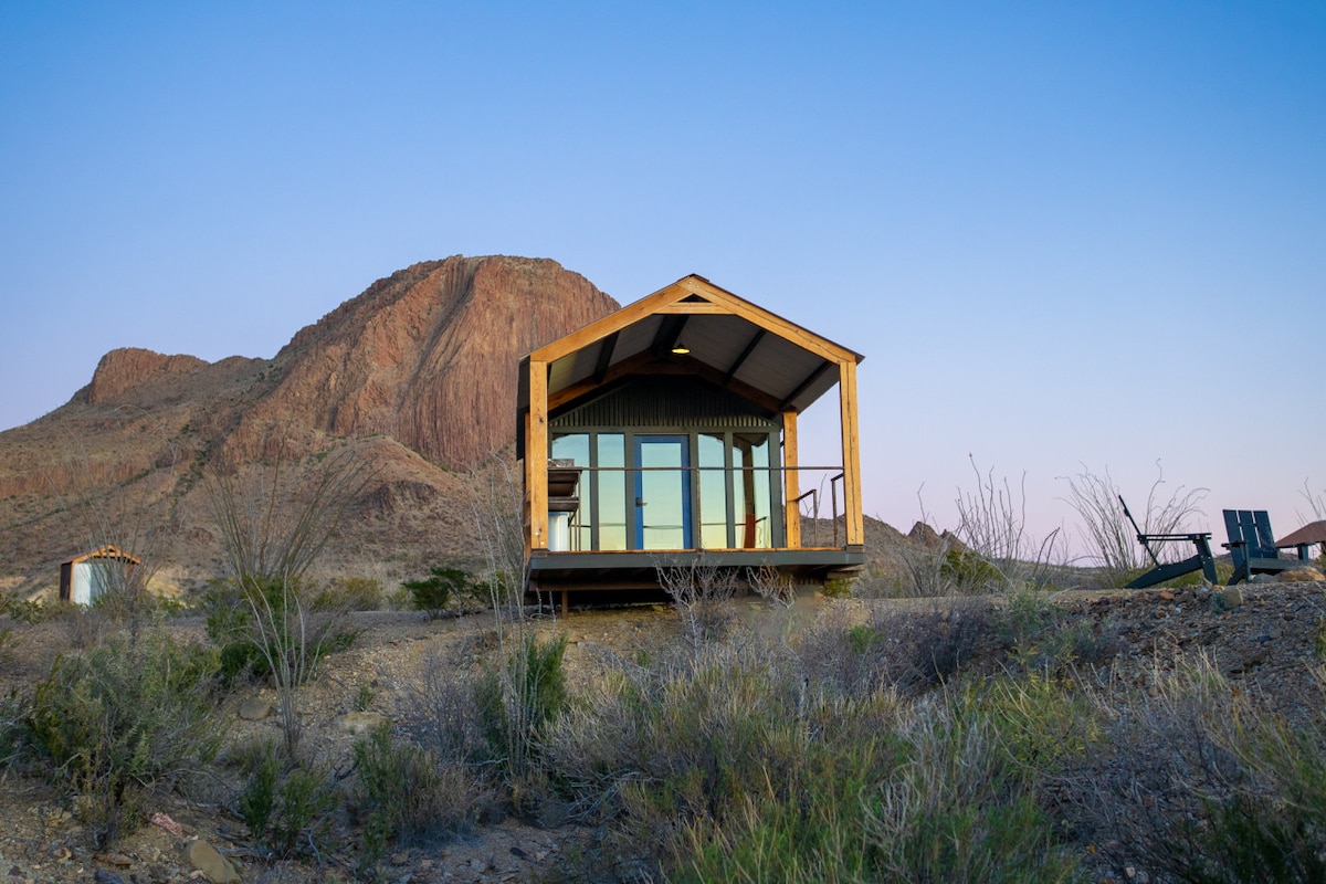 A rustic modern cabin is positioned against a dramatic mountain backdrop, featuring large glass windows that reflect the natural landscape. The elevated deck offers a spacious area for relaxation, surrounded by desert vegetation, with a soft pastel sky above as day transitions to evening.