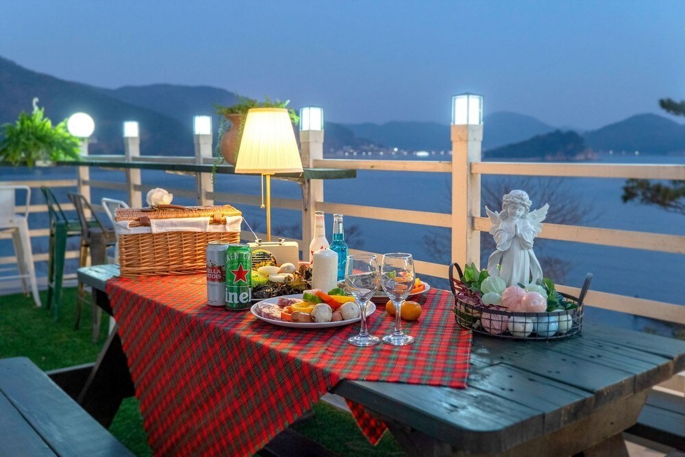 An outdoor dining area is set with a checkered tablecloth, featuring a picnic basket, sparkling glasses, and a variety of snacks. Soft lighting from a lamp enhances the serene atmosphere, while scenic ocean views and distant hills create a calming backdrop.